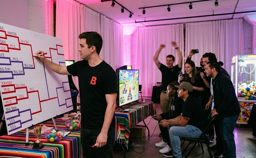 A Budcade host updates a tournament bracket while players race and friends cheer at a game night